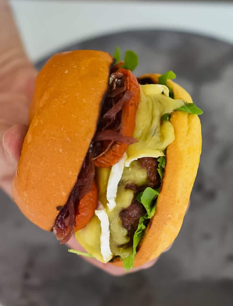 Organic Beef Burger someone holding an organic beef burger with brie, roasted tomatoes & onion marmalade