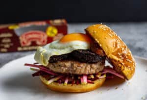 Pork & black pudding burger on a white plate with top bun off. Clonakilty pudding packet in background