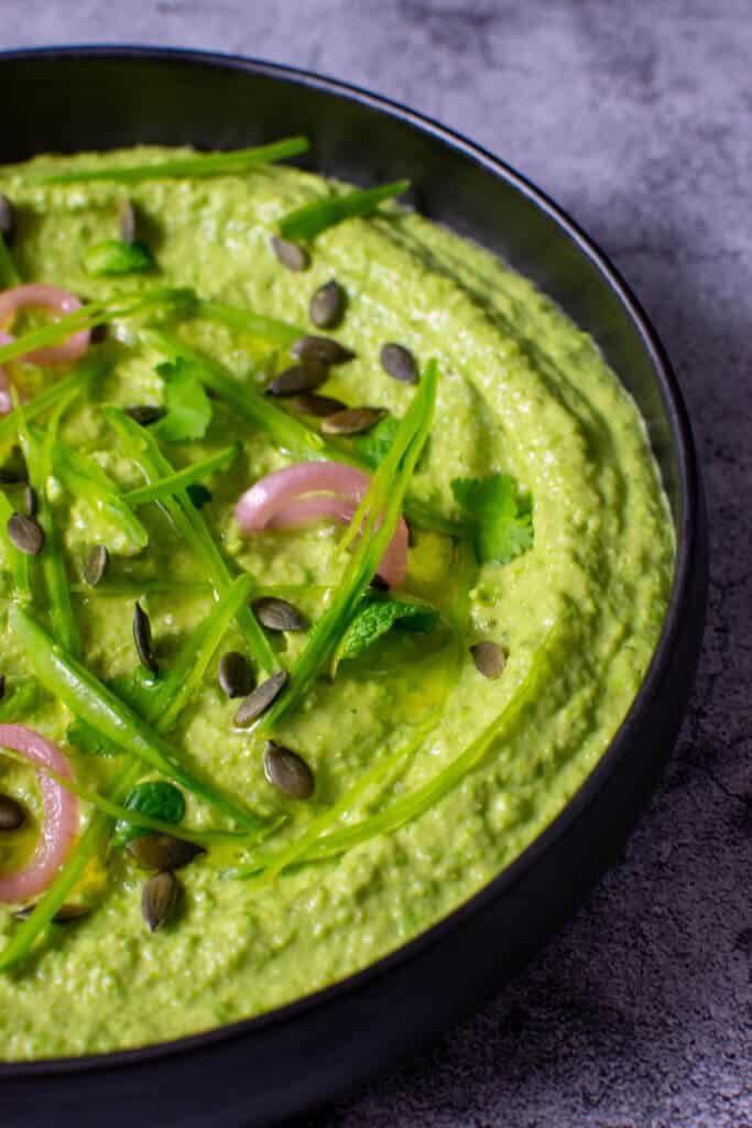 black bowl with avocado & pea hummus topped with pickled onions, pepitas, herbs & snowpeas