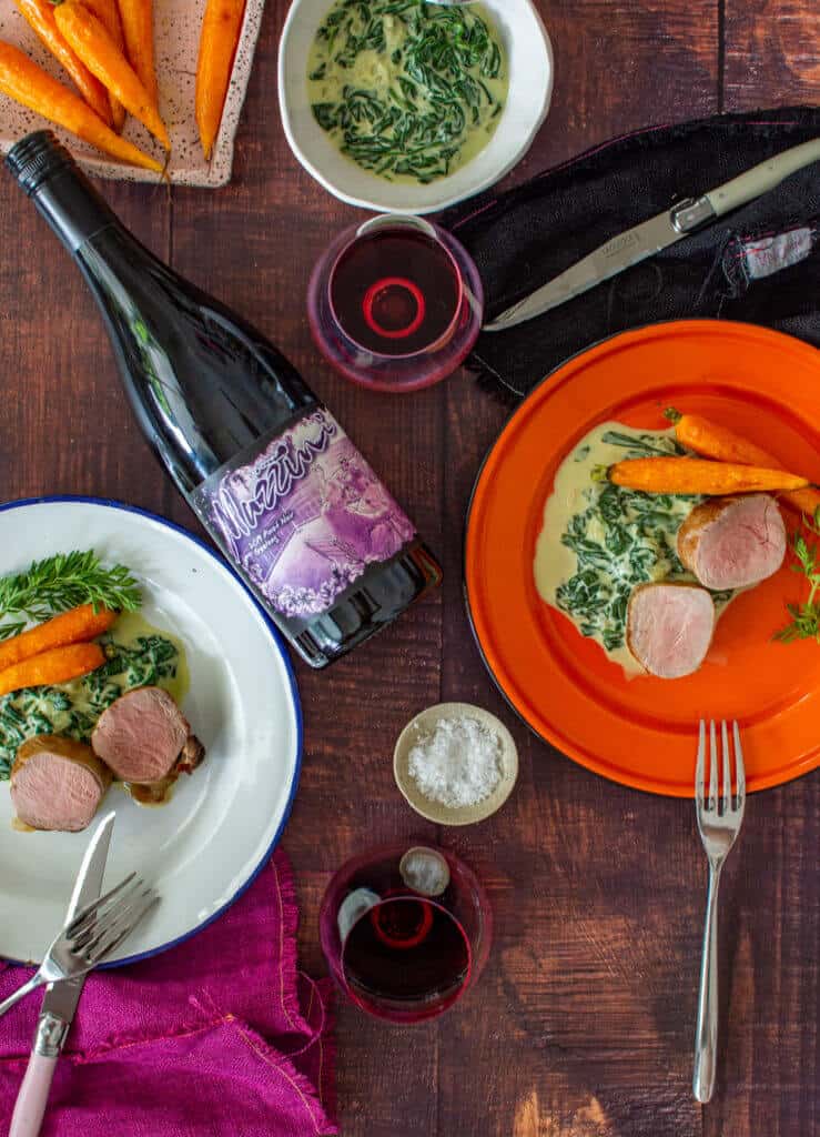 2 plates of pork tenderloin, creamed spinach & carrots, mazzini pinot noir and cutlery on a table