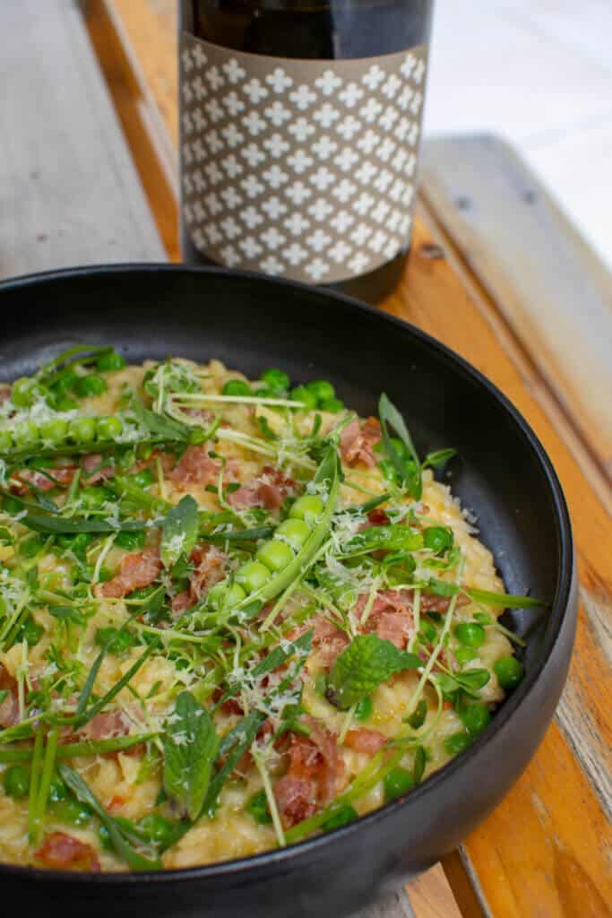 pea & bacon risotto in a black bowl on wooden table with heirloom chardonnay
