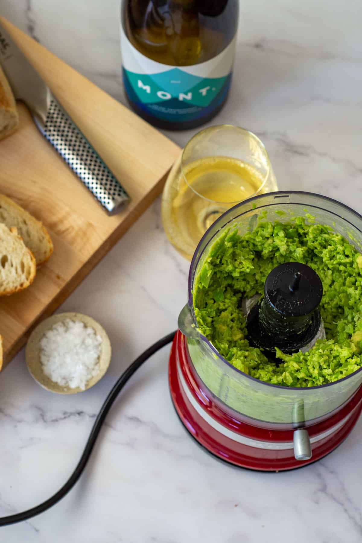 crushed edamame & avocado in a blender with mont wines chardonnay in background and bread on a chopping board
