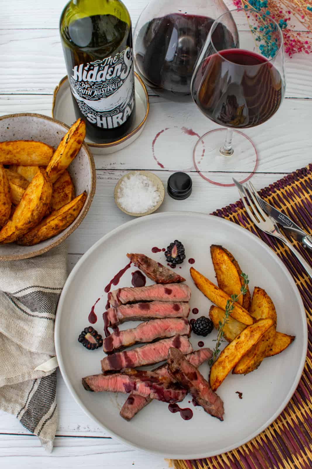 cooked scotch fillet, spicy potato wedges and blackberry jus on a plate with bowl of potato wedges and bottle of hidden sea shiraz next to it