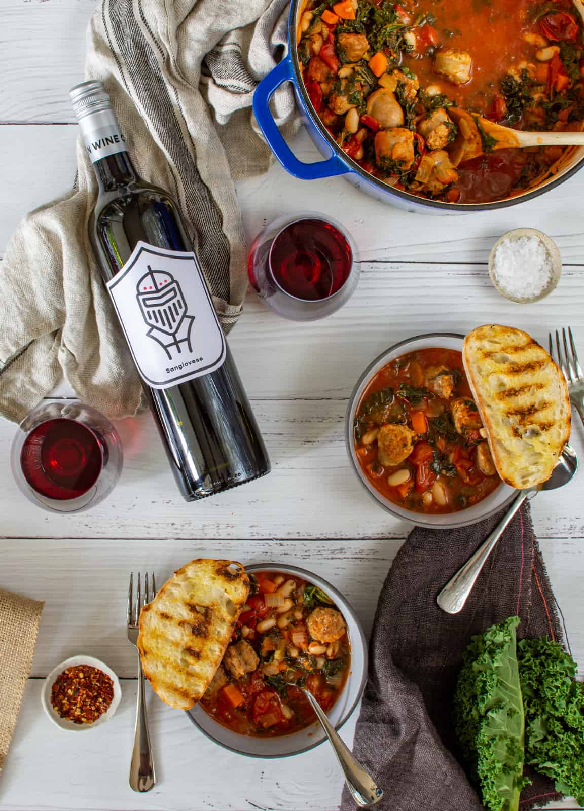 tuscan sausage stew in blue casserole dish, bowls of stew and pawn wine co red wine on a white table