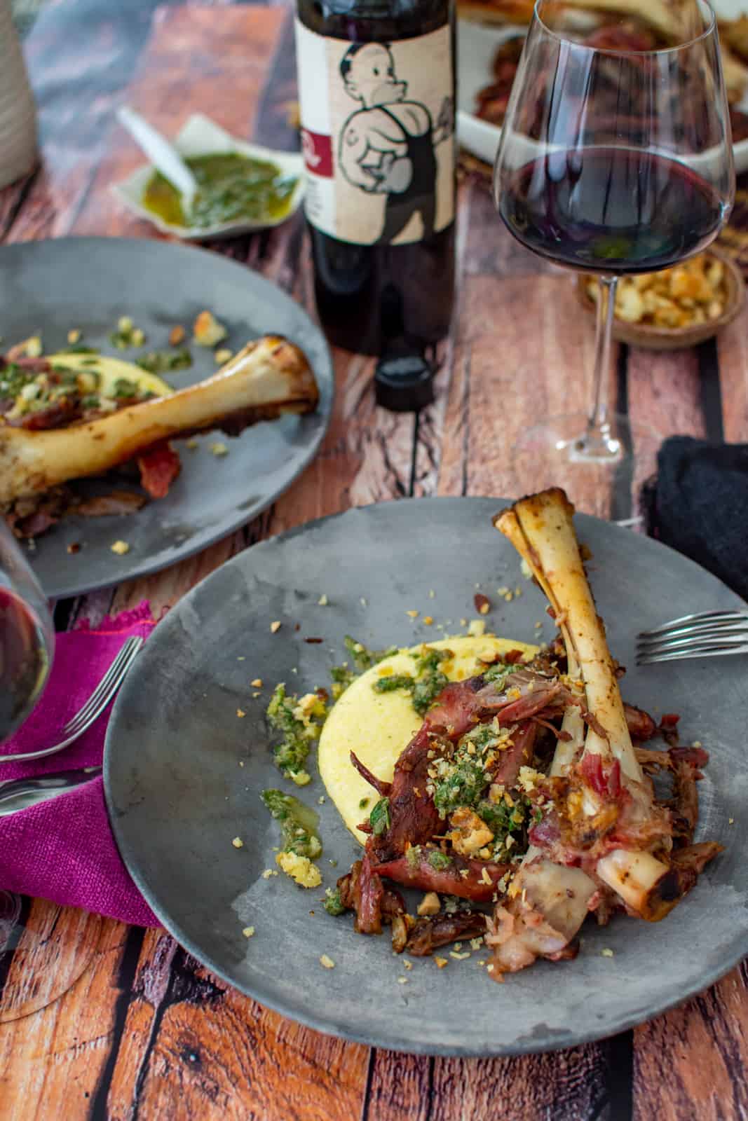 smoked lamb shanks on a plate with polenta and anchovy dressing, wine glass and mollydooker wine in background