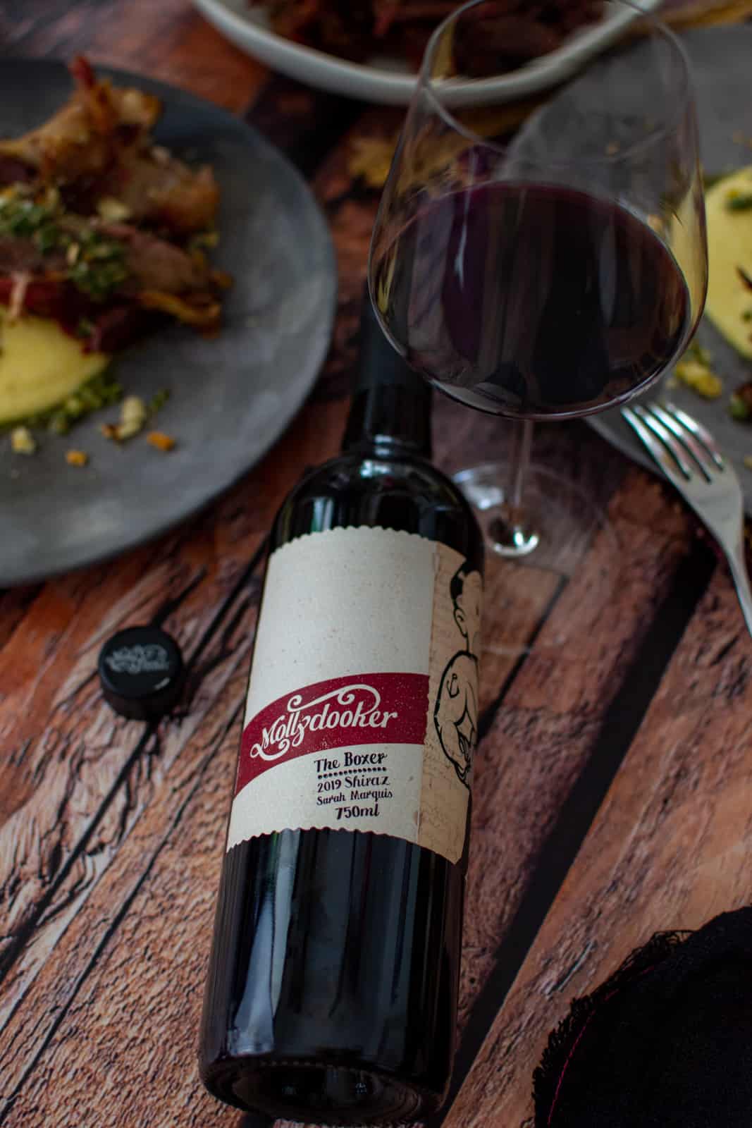mollydooker wine lying on a table, wine glass and smoked lamb shanks on a plate in background