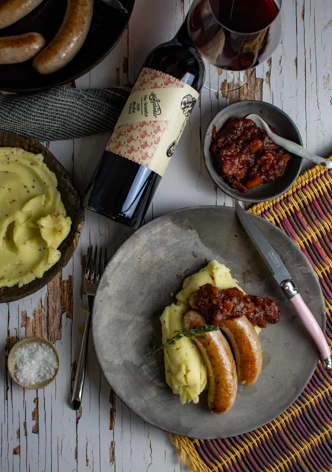 gourmet bangers and mash on a plate with mashed potatoes in a bowl, cutlery and red wine