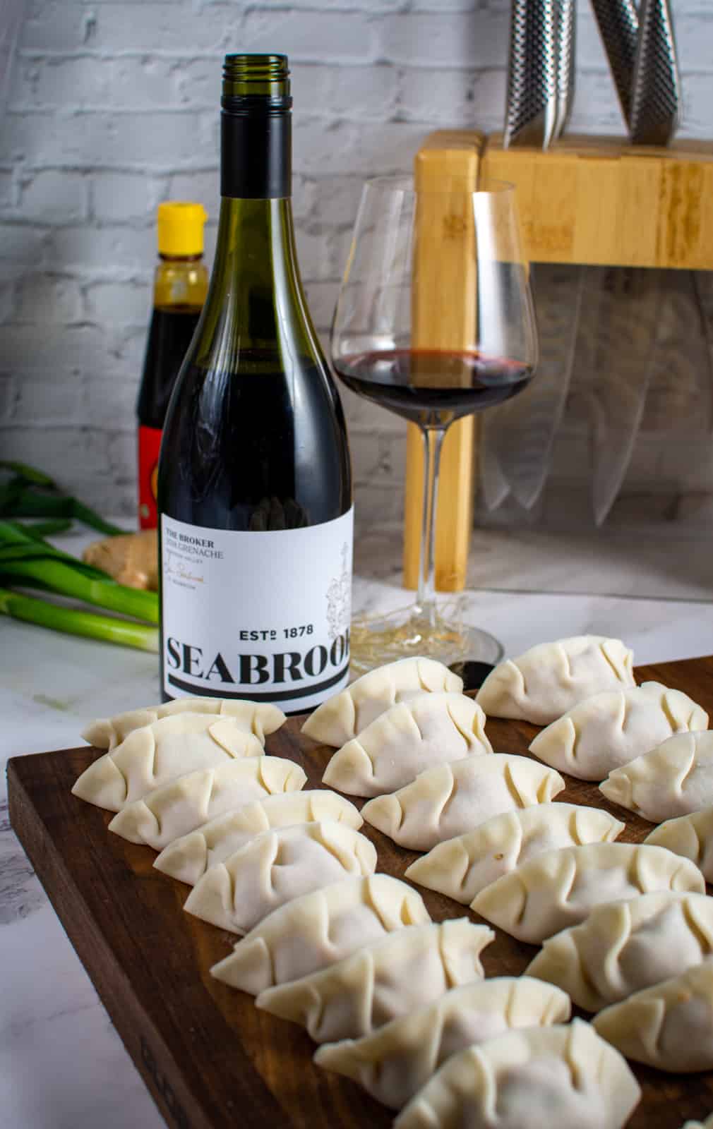 a chopping board with multiple pork cabbage dumplings and bottle of wine in background