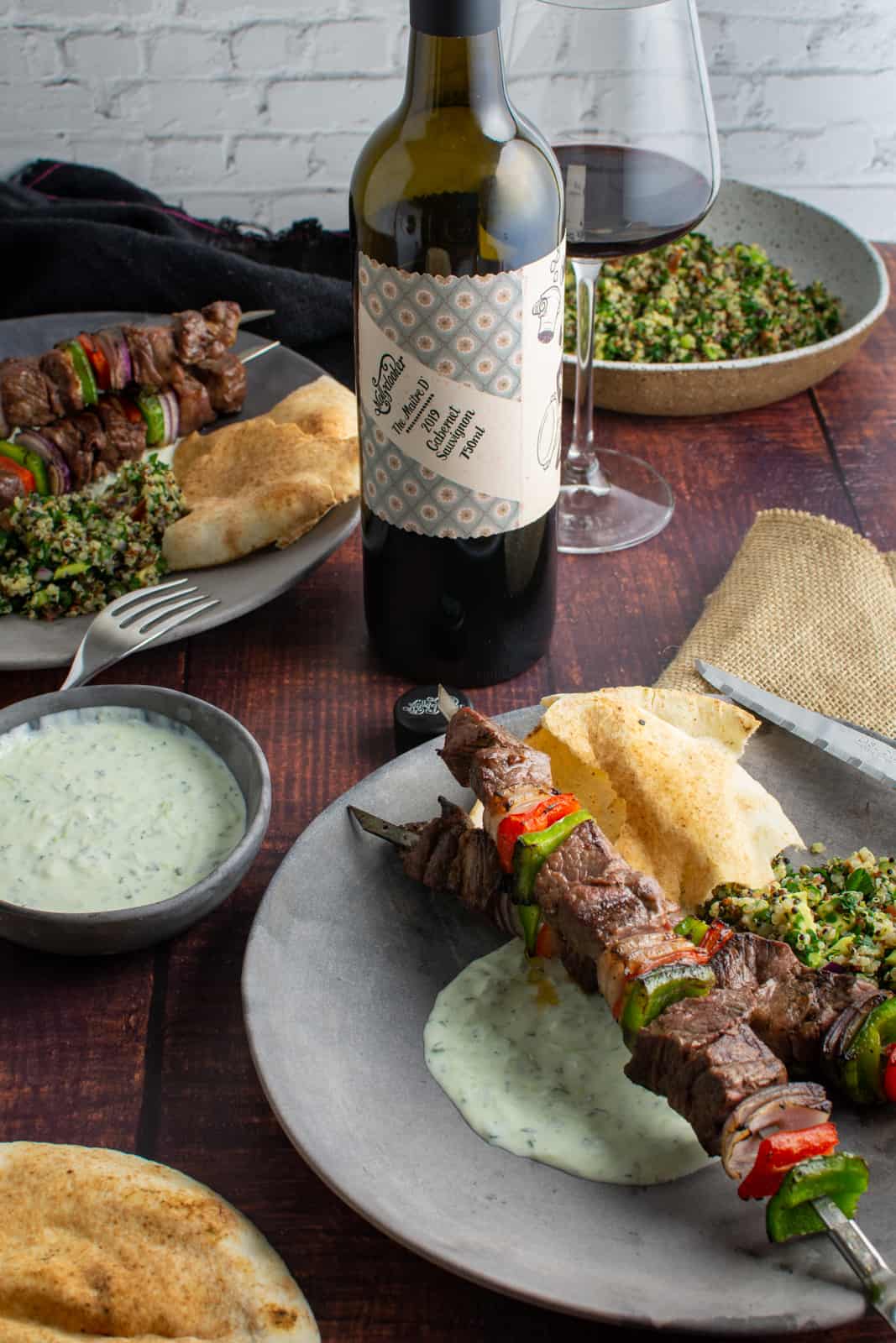 grilled lamb kebabs on a plate with yoghurt dressing, pita and red wine in backgrund