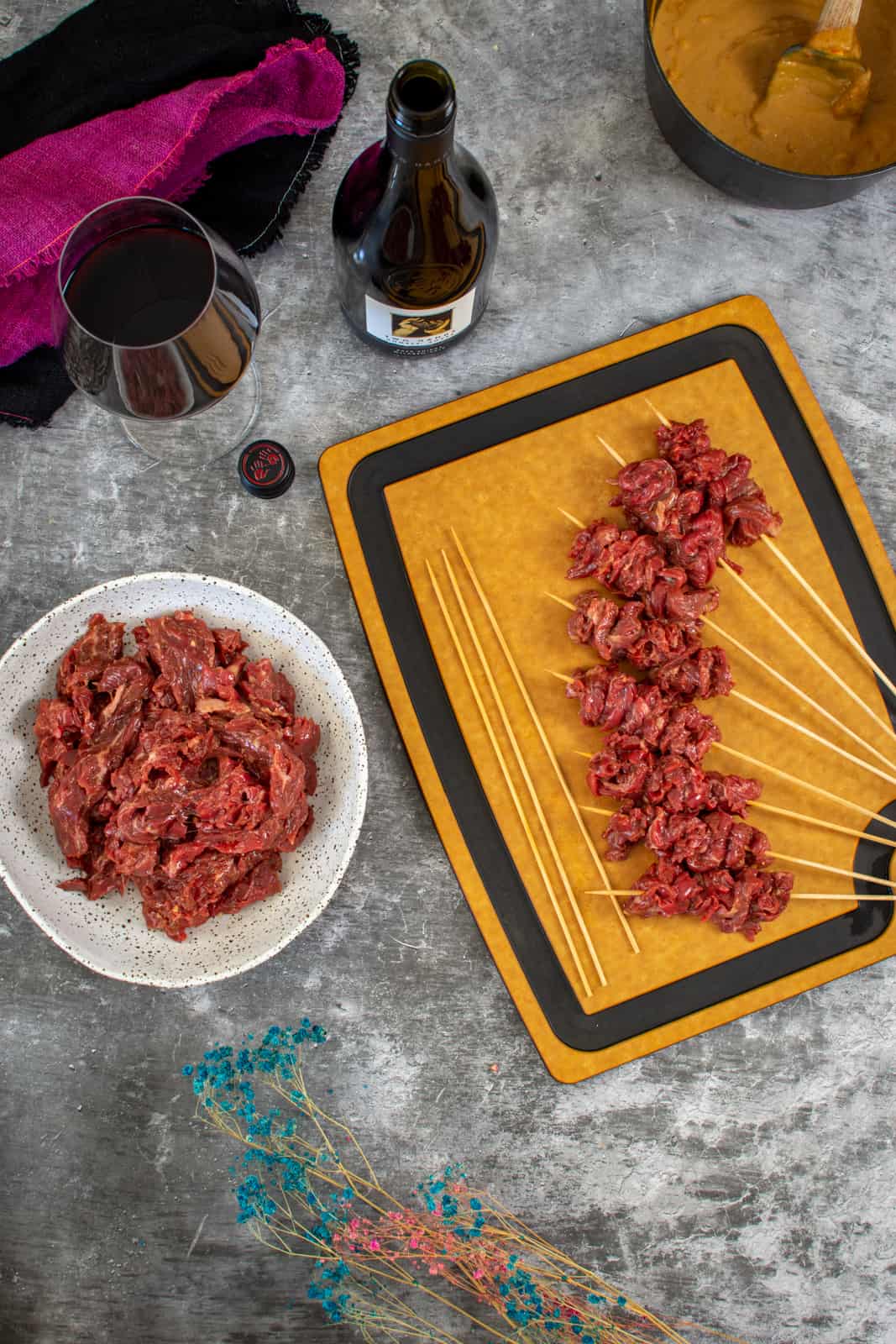 beef skewers on a chopping board with bowl of marinated beef and wine next to them