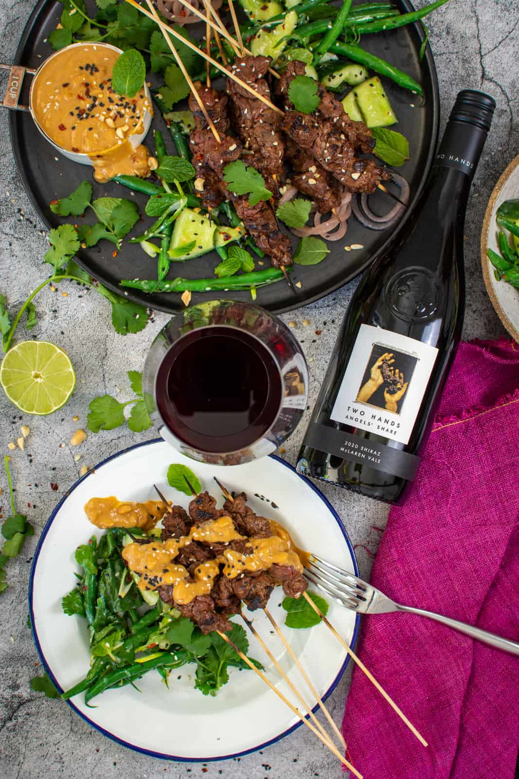 beef satay skewers and cucumber salad on a plate, red wine bottle next to them 