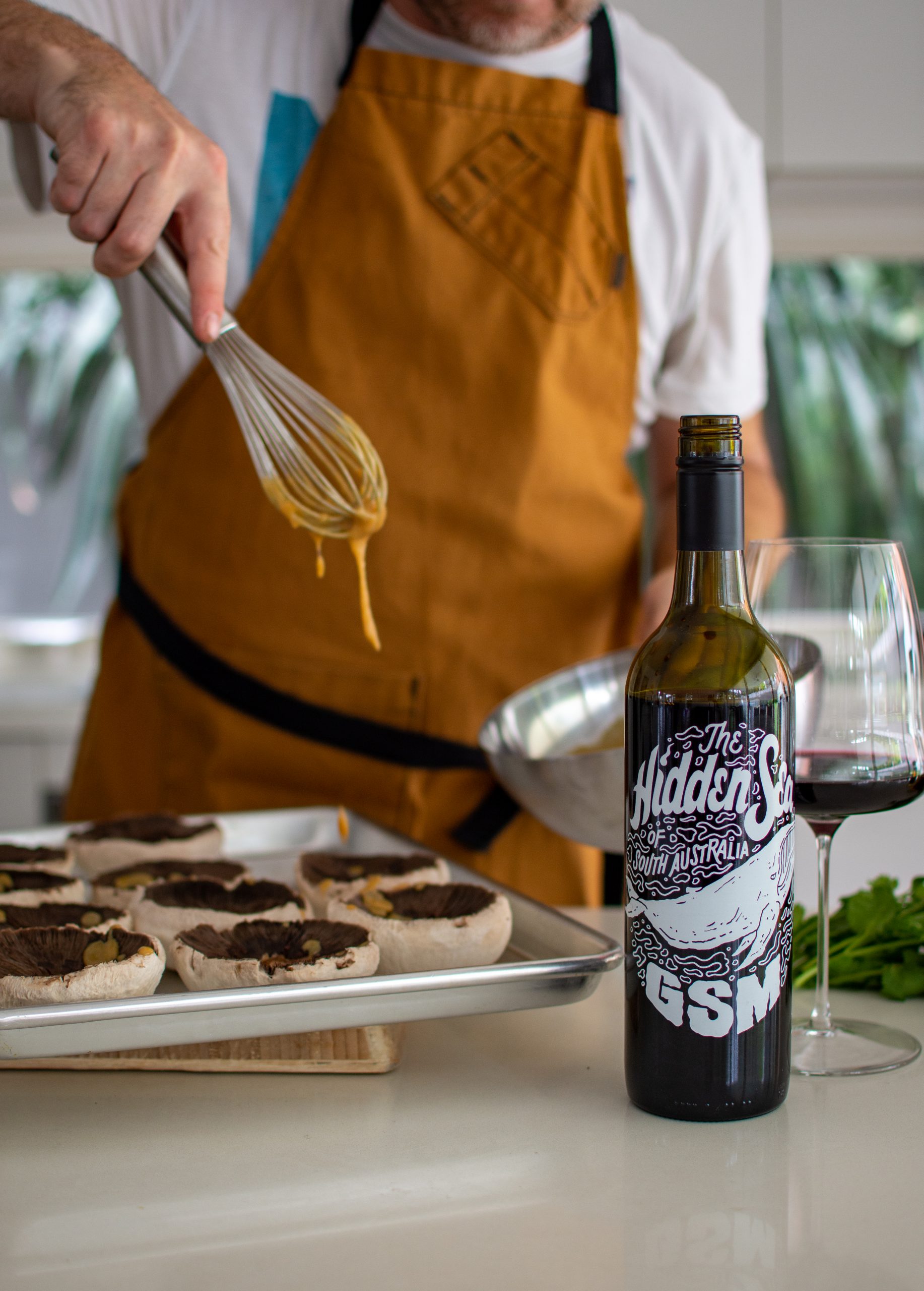 Someone drizzlng miso dressing over mushrooms in the kitchen with food and wine on counter top
