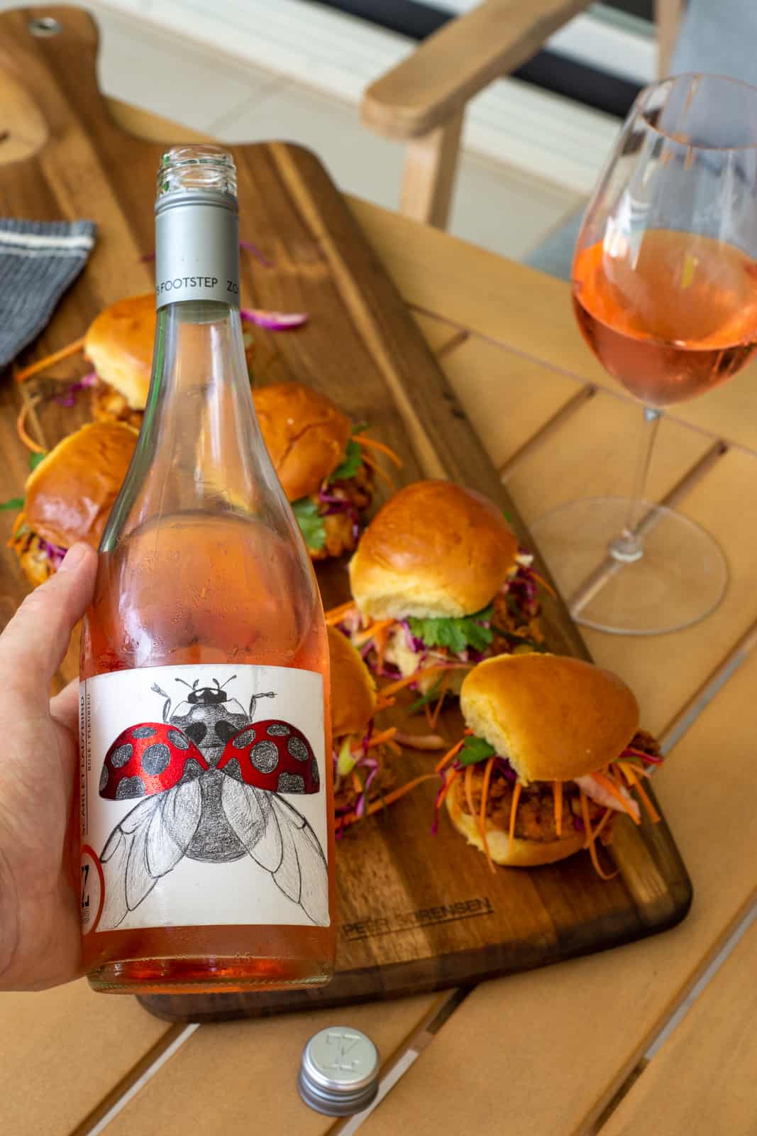 someone holding a bottle of rose wine above a board of fried chicken sliders