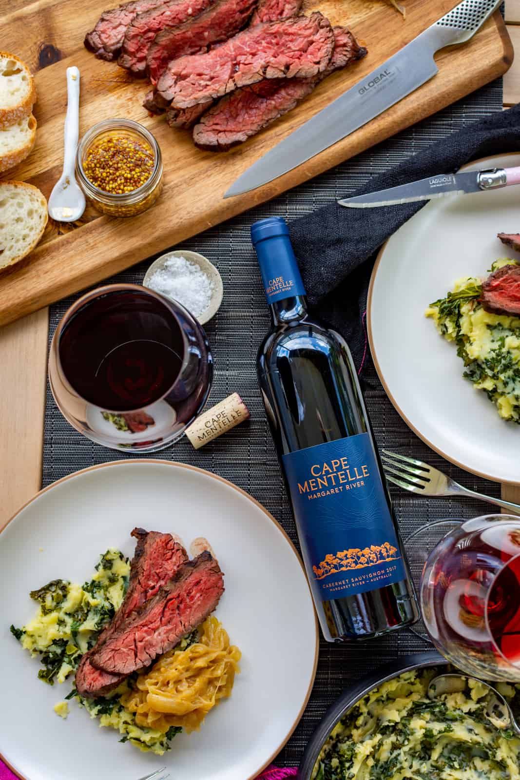 birdseye view of plates with miso steak and colcannon mash. Red wine and glasses beside them on table