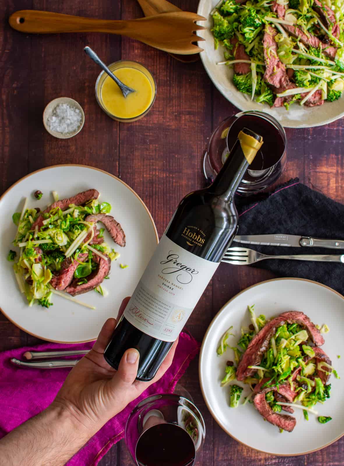 someone pouring red wine with broccoli, steak & pear salad on plates below