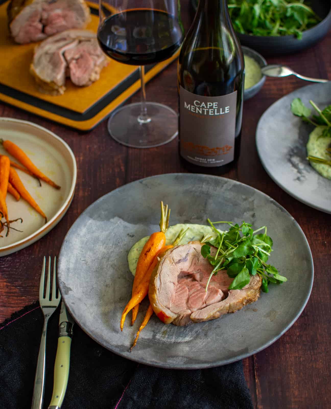 roast lamb on a plate with carrots and chermoula yoghurt. wine and roast lamb in background