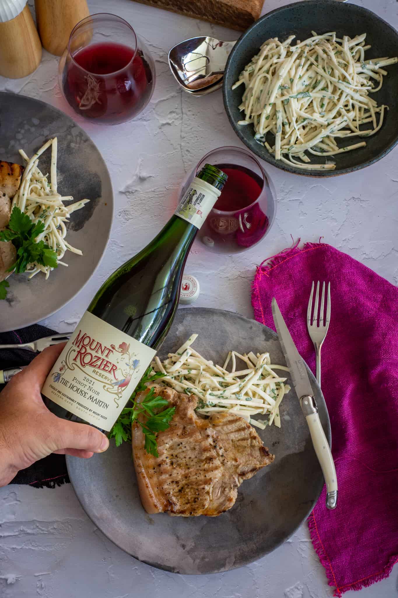 Someone pouring pinot noir with plates of grilled pork & celeriac remoulade below