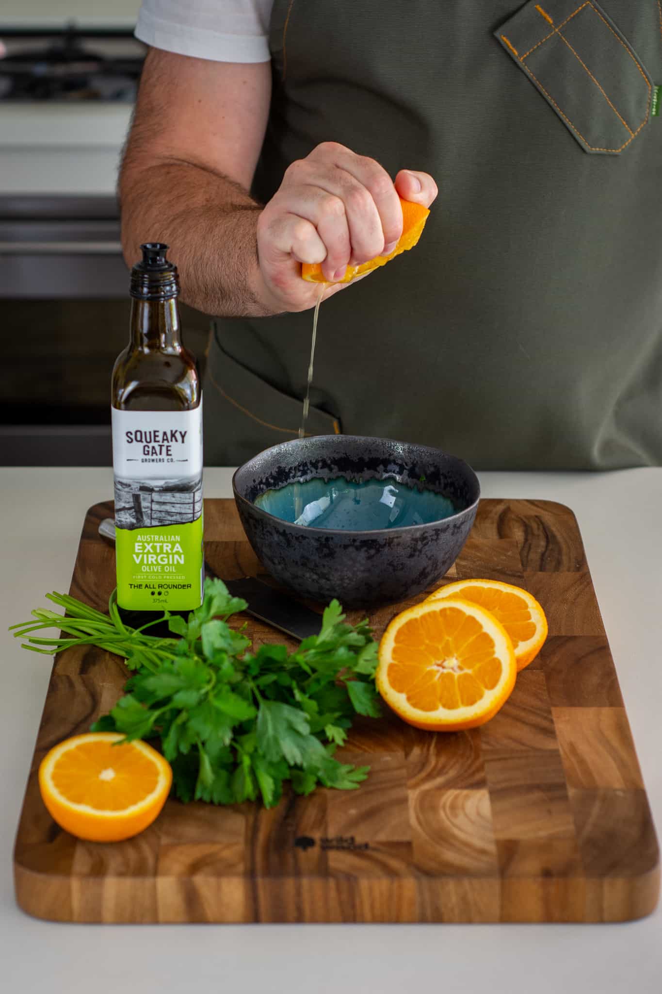 Someone squeezing a orange into a bowl with parsley and olive oil on a chopping board beside the bowl