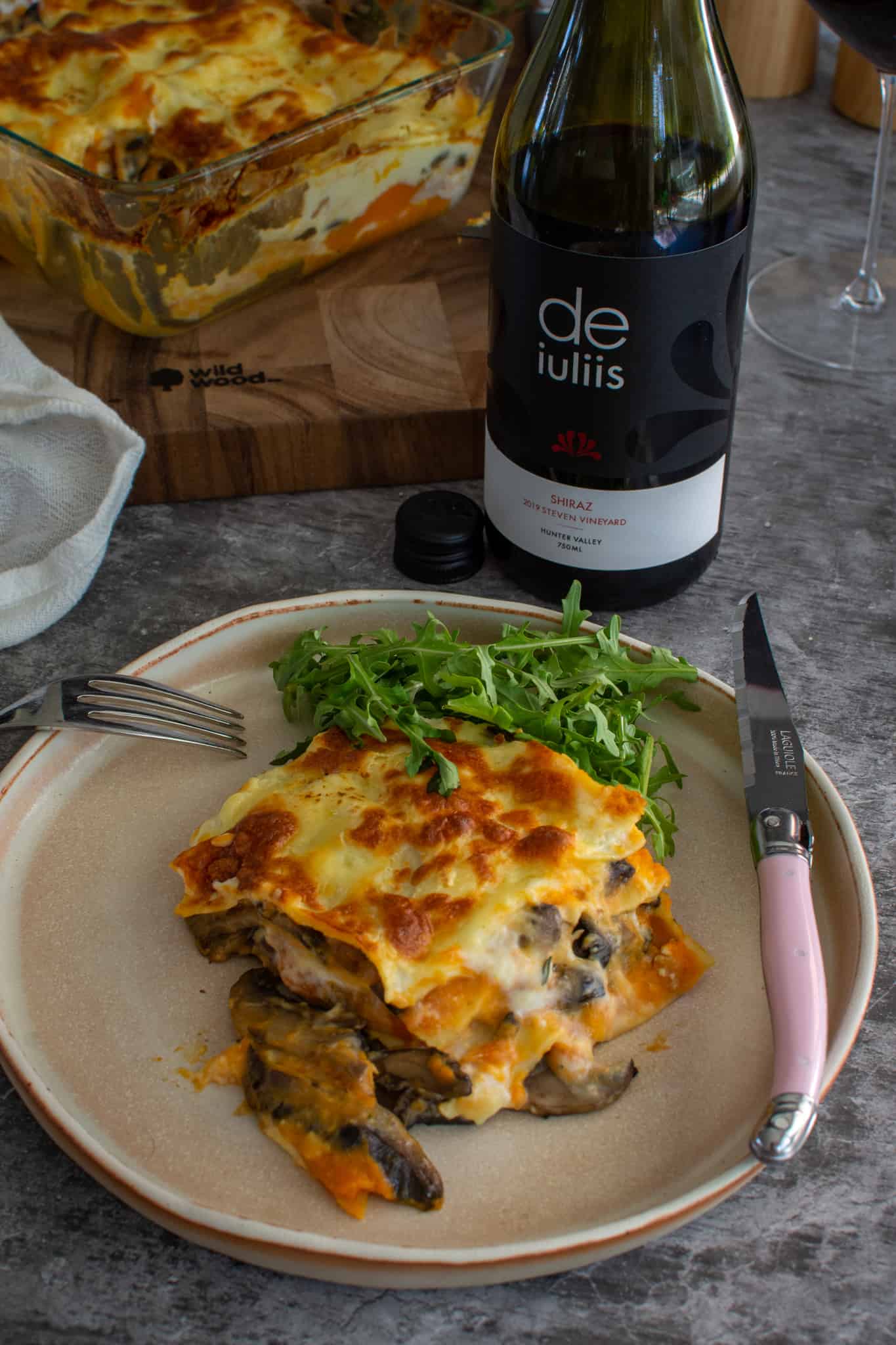 Vegetable lasagne on a plate with a small bit of rocket salad too. De iuliis wine beside it with lasagne dish in background