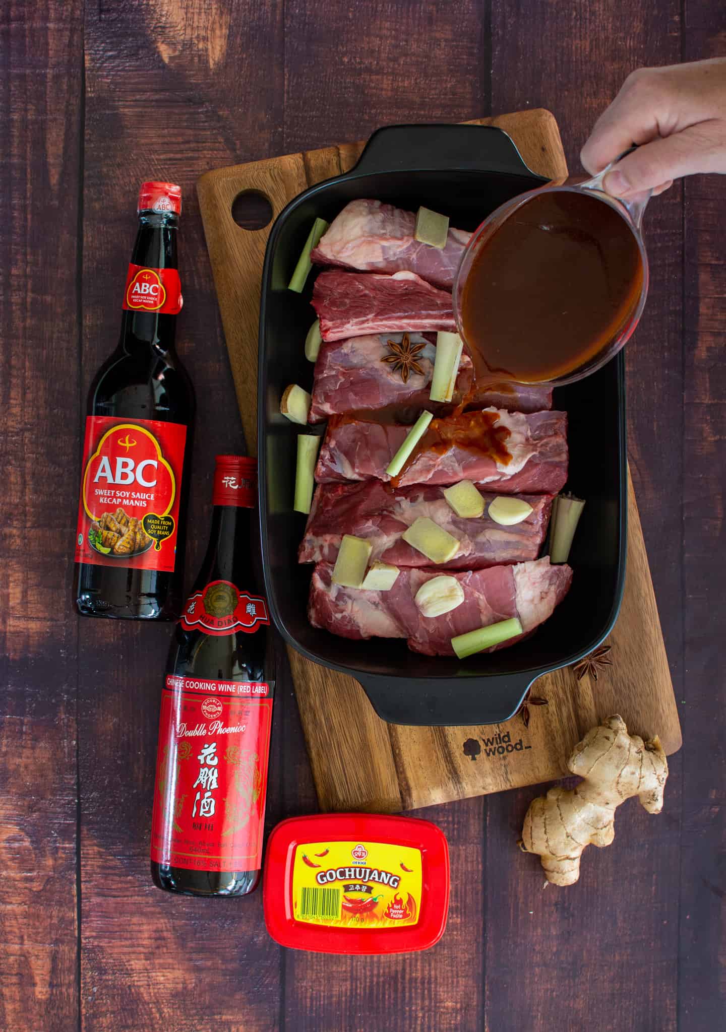 someone pouring braising liquid into a pot of beef short ribs with Asian ingredients beside the pot