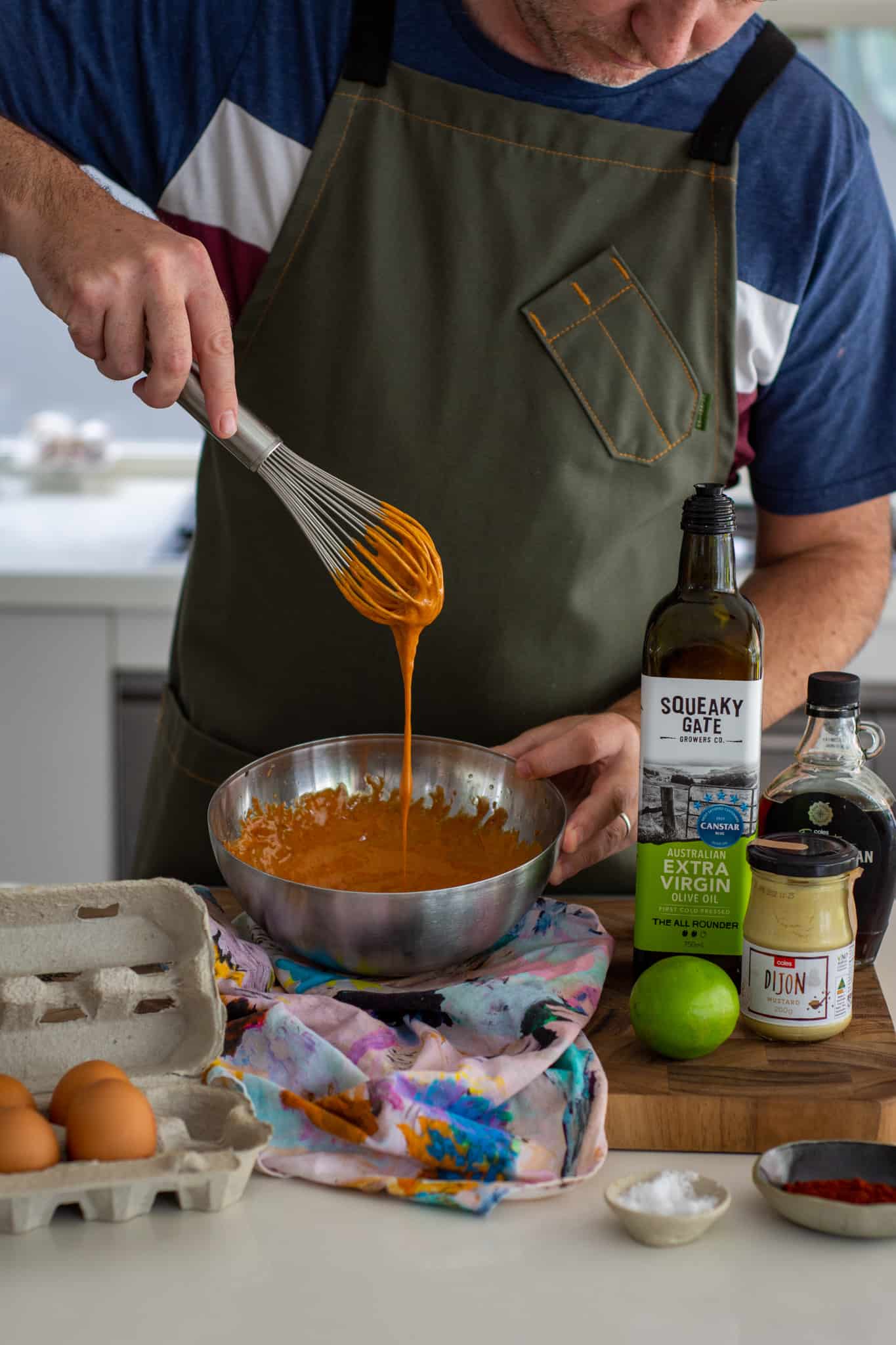 someone making smoked paprika aioli by hand. the aioli is falling off a whisk into a bowl with ingredients on counter top