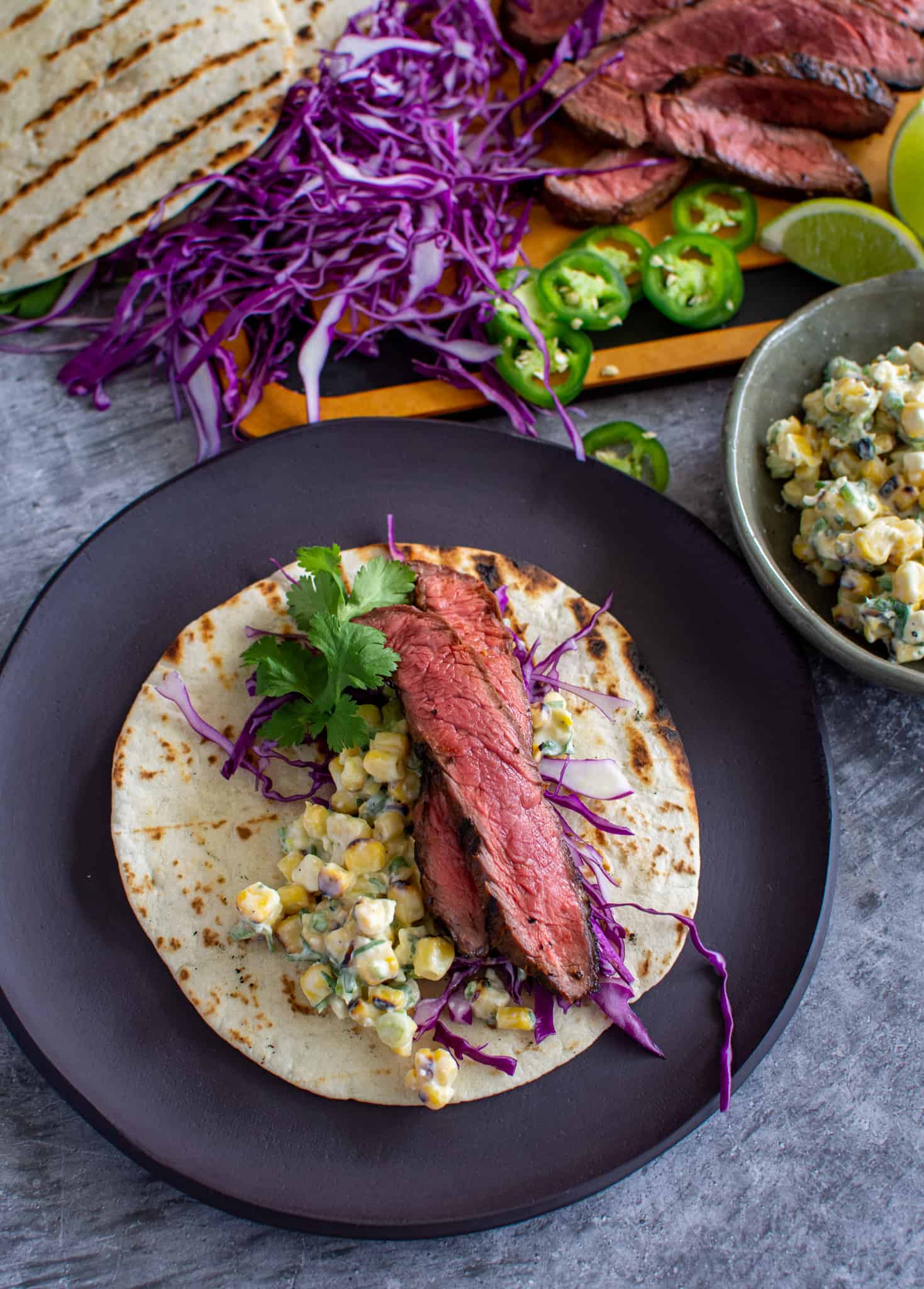 close up of 1 steak taco with corn elote with sliced steak, red cabbage and tortillas in background  