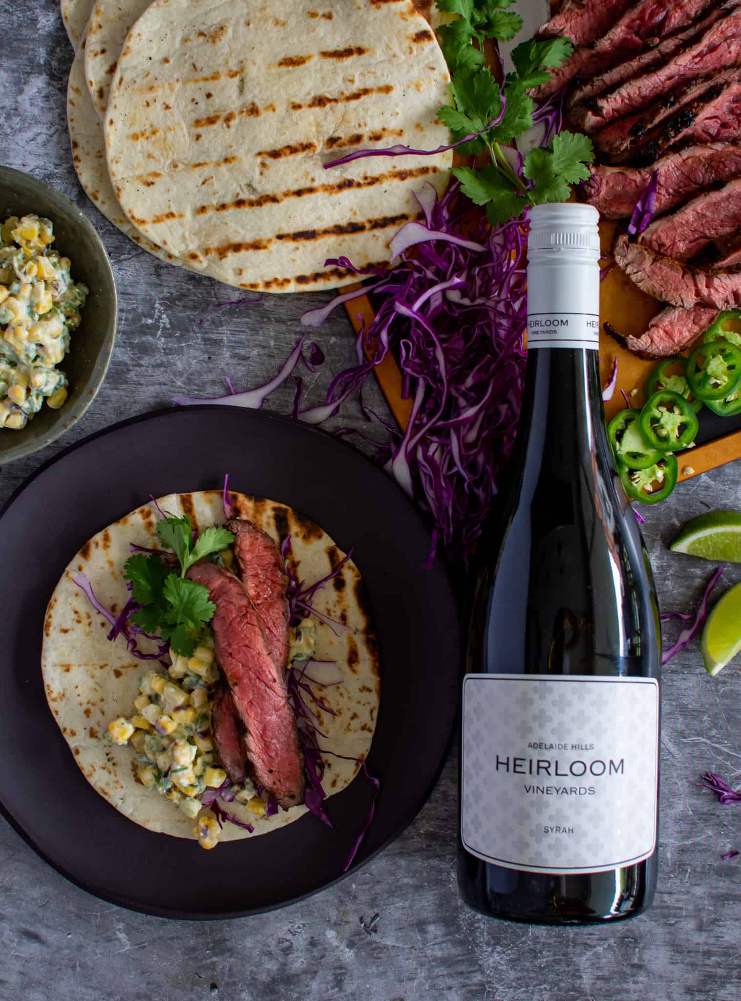 Birdseye view of grilled steak tacos with red cabbage, corn elote and a bottle of Heirloom Syrah next to them