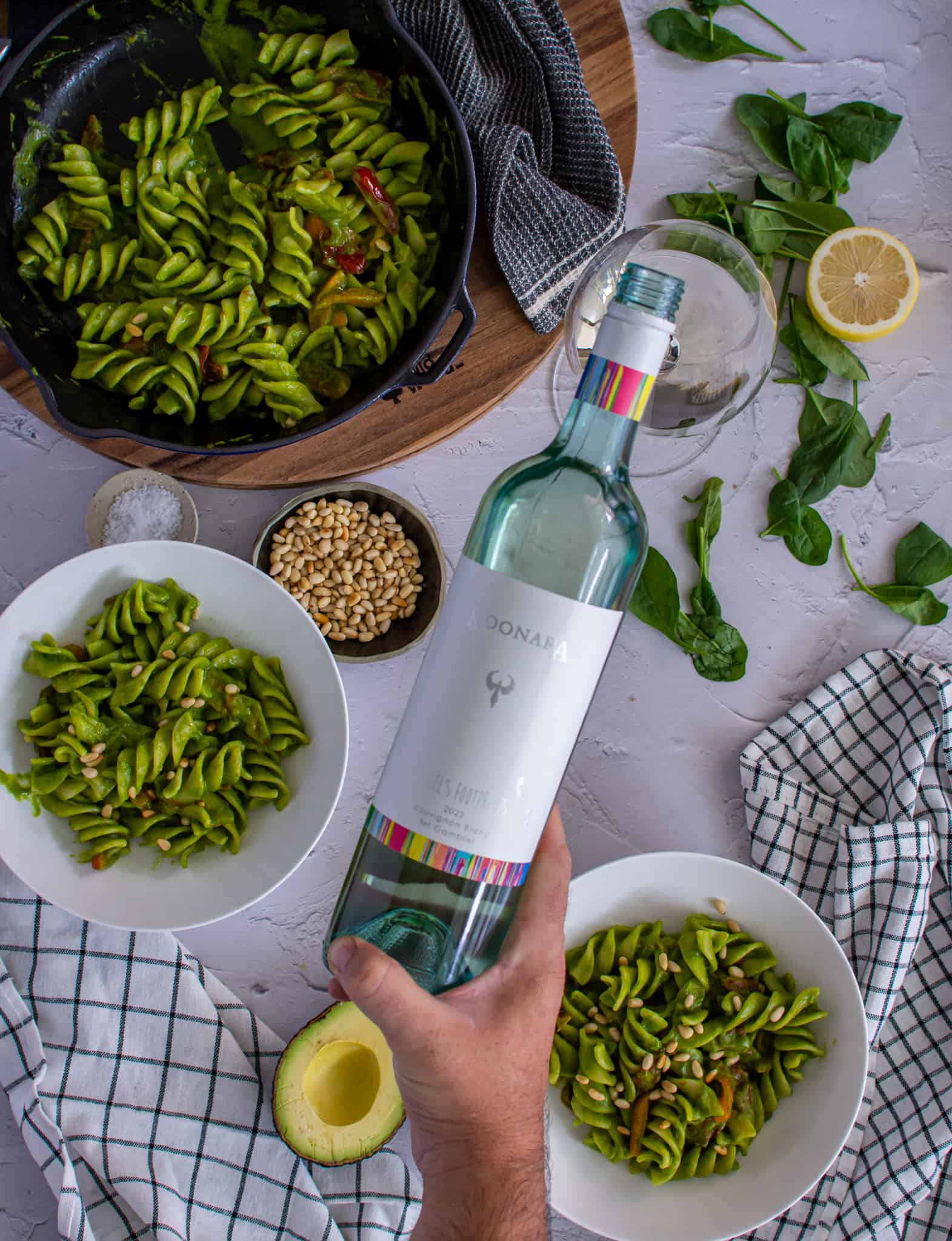 birdseye view of avocado pasta in saucepan and bowls of avocado pasta. Someone pouring a glass of white wine too
