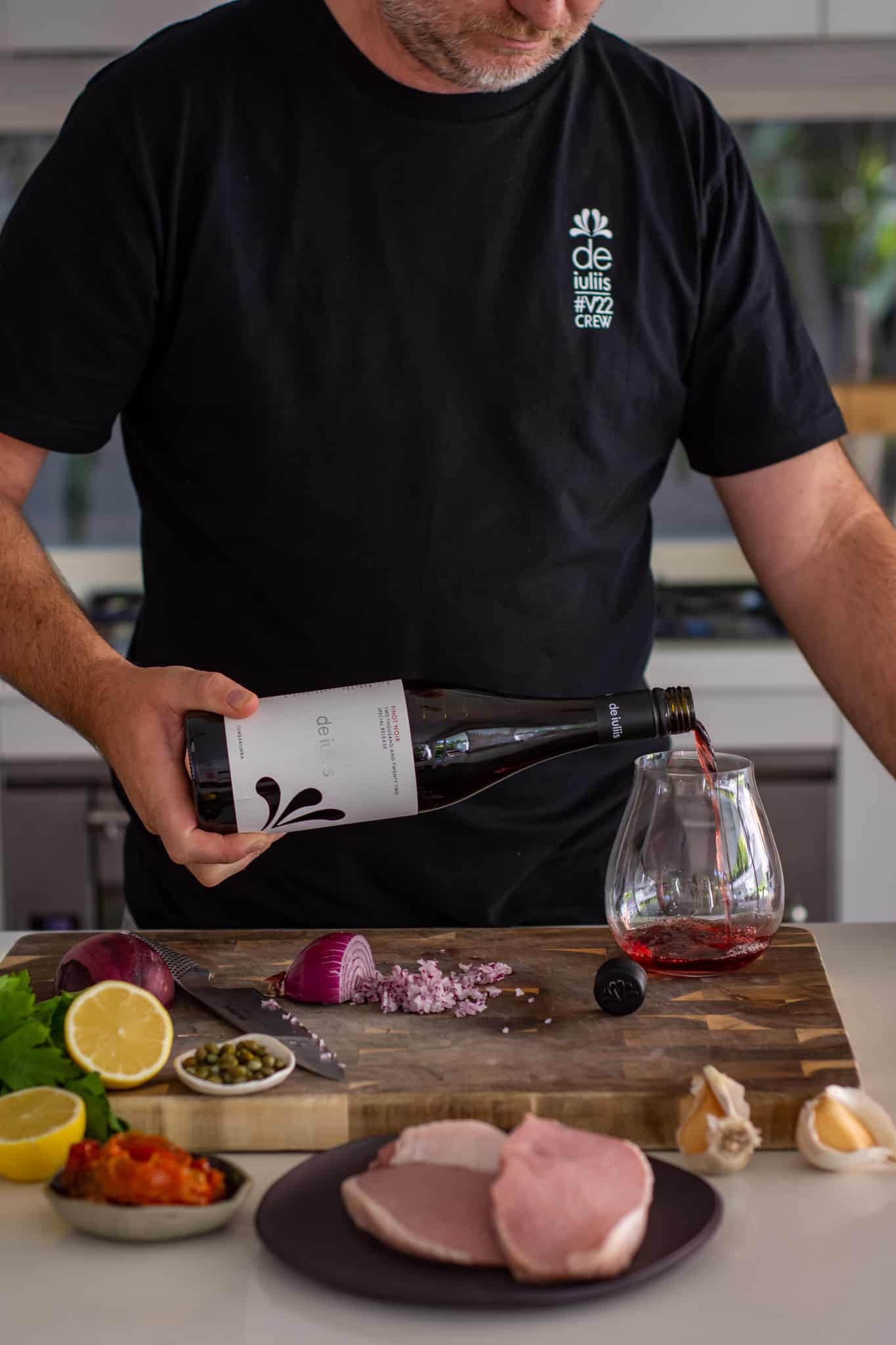 someone pouring red wine into a glass in the kitchen with ingredients on chopping board