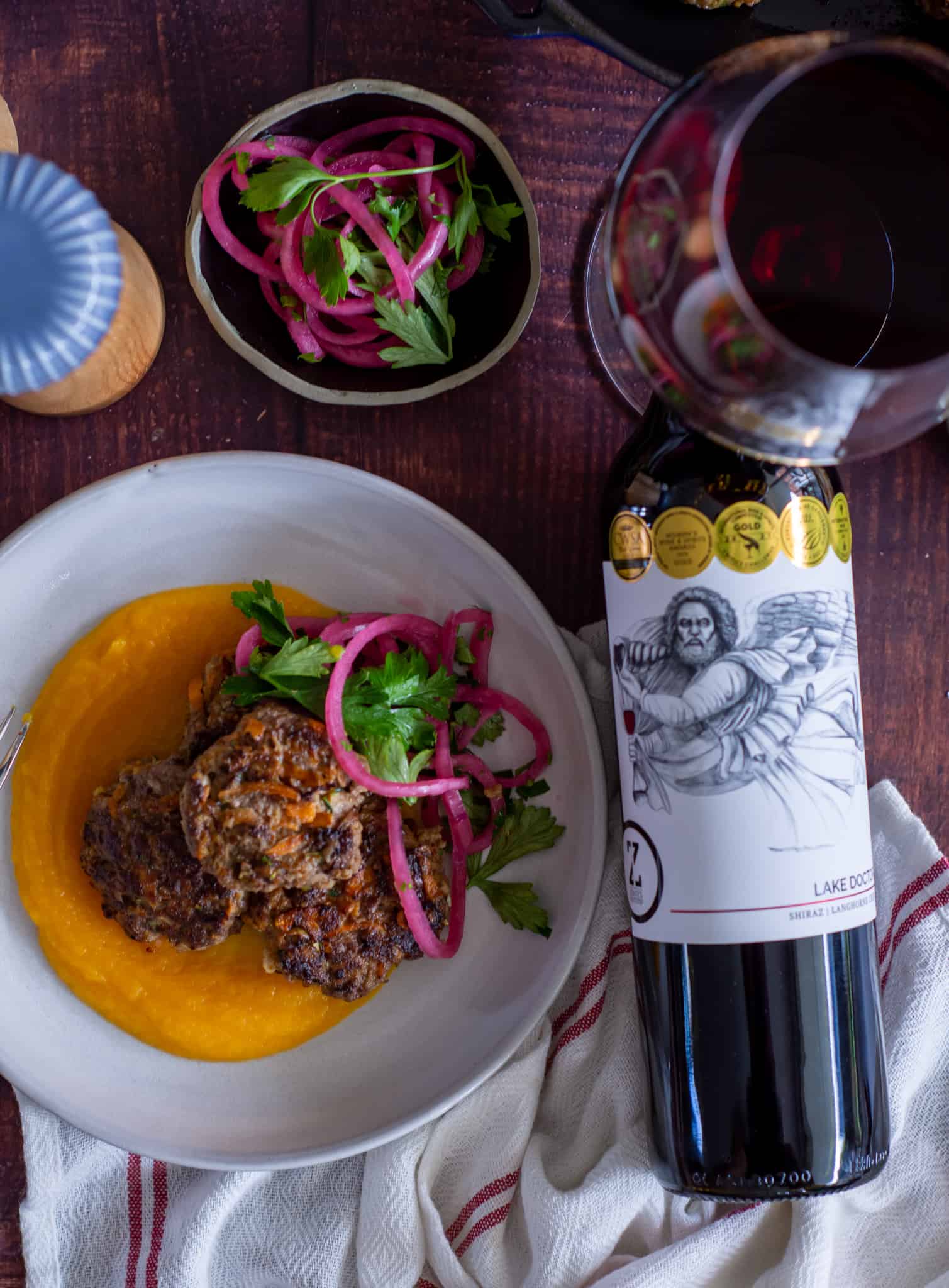 birdseye view of rissoles on a plate with bottle of wine beside plate, wine glass and also bowl of pickled onions